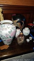 Overview of various vases, jars, and cups on wood surface including large floral vase, black vase with crane, lidded jars, glass dome jar, and small cups