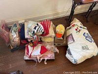 Wide view showing the stacked vintage baby clothes, folded quilt, and large plush duck head in a box.