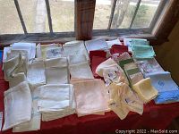 Photo of various folded vintage table linens and napkins in multiple colors including white, red, green, blue, and patterned, laid out on a red surface near window light.
