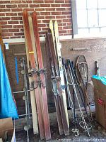 Overview of vintage ski equipment including wooden skis, snowshoes, lacrosse racquet, and ski poles leaning against wooden panel and brick wall in garage.