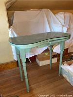 Full front angled view of green painted vanity with curved glass top, placed on wooden floor with background items covered in white sheets.