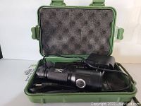 Photo of a black rechargeable flashlight with textured grip, power button, charging cable, and wall adapter inside an open green protective hard case with foam padding.