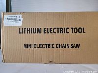 Cardboard box with printed text 'LITHIUM ELECTRIC TOOL MINI ELECTRIC CHAIN SAW' indicating a mini electric chain saw powered by lithium battery.