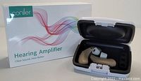 Photo of two beige Conlier hearing amplifiers with white earbuds in a white charging case, next to the original product box with colorful wave design and product name.