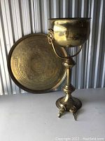 Photo showing the large decorative round brass platter and pedestal urn in front of a corrugated metal background.