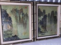 Two framed Chinese landscape paintings with bamboo-style frames leaning side by side, showing traditional scenes of mountains and water. Frames and glass show visible scratches and dust.
