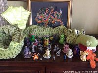 Full view of large stuffed green dragon with multiple dragon figurines arranged in front and framed needlepoint dragon artwork in the background.