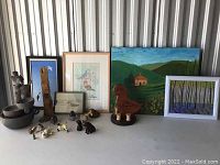 Photo showing various framed artworks depicting nature scenes and birds, two stacked stone birdbaths with bird sculptures, wooden carvings, and multiple small animal figurines arranged on a table