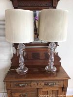 Pair of glass table lamps with pleated white fabric shades on wooden table in front of wooden mirror.