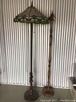 Full view of Tiffany styled stained glass floor lamp with wooden base standing next to separate wooden floor stand.