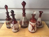 Photo showing five vintage glass decanters of various shapes, decorative bird and floral motifs in amber/red and white colors, placed on a wooden surface.