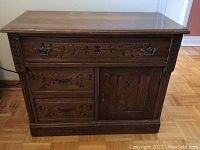 Full front view of the wood cabinet showing three drawers and a door with carved floral and leaf decorations, dark wood finish.