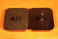 Two black Apple TV devices side by side on a wooden surface, showing signs of wear and tear.