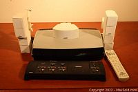 Bose stereo system components showing two white cube speakers, black media center, video enhancer, and remote control on brown surface.