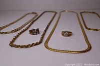Four gold plated necklaces arranged side by side with two gold plated rings in front. Necklaces vary in chain style and thickness. Rings include one with multiple rhinestones and one with a single large rhinestone.