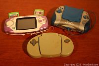 Three handheld electronic game devices displayed on wooden surface: purple and white Leapster with cartridges, blue and silver Leapster L-Max, and beige 'Computer Games Calculator' game.