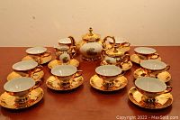 Full tea set arranged on table including 8 cups and saucers, teapot, creamer, and sugar bowl, all gold plated with matching romantic scene artwork