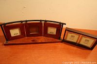 Two photo frames: one three-panel connected frame and one double frame, both wooden with black metal support or stand, sitting on wooden surface against a plain wall.