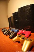 Four black Sony PlayStation 3 consoles standing upright in a row with seven wireless controllers laid out in front, capturing all items included in the lot.