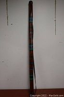 Full length view of the wooden rain stick with decorative painted bands and carrying strap hanging on wall.