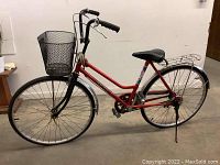 Full side view of red bicycle showing frame, front basket, rear rack, pedals, kickstand.