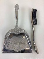 Front view of the ornate silver table sweep along with the brush showing decorative handles and detailed engraving.