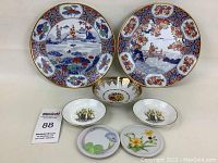 Seven assorted ceramic dishes arranged to show designs and relative size, including scenic plates, floral bowls, and small plates