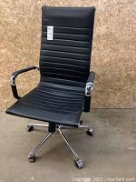 Front view of black vinyl office chair with padded armrests and metal base with casters on concrete floor and wood background.