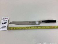 Side view of unused Goya bread knife showing serrated blade and black handle, a measuring tape shows approx 28 cm blade length.
