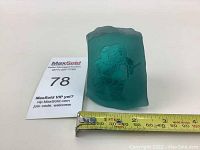 Frosted teal glass sculpture offset with an Inuit figure etched on the front, measuring about 3 inches wide, shown with a tape measure for scale.