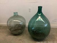 Two large vintage glass jars placed side by side on the floor against a wall. The jar on the left is clear and round. The jar on the right is green and teardrop shaped.