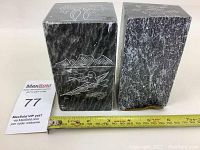 Two dark soapstone block carvings side by side with a measuring tape showing approximately 12 cm height and a MaxSold lot number card.