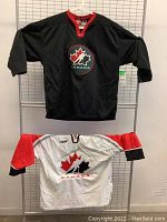 Two Team Canada ice hockey jerseys, one black and one white with red and black sleeves, hanging displayed together.