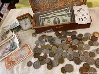 Wide view of assortment coins and banknotes laid on white cloth with a wooden display box holding US banknotes and auction tag seen
