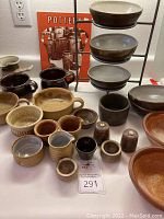 Wide view of entire assortment showing various pottery pieces including mugs, bowls, sugar bowl, shakers and pitcher arranged on table with pottery catalog in background.