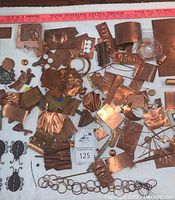 Photo of various copper and brass scraps including multiple shaped sheets, discs, wire coils, and metal loops spread on white background with a red measuring stick for scale.