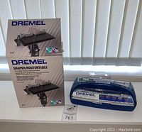 Two Dremel Shaper/Router Table boxes showing front and side views, placed next to a blue Dremel accessory bit kit case on a white surface with vertical blinds in the background.