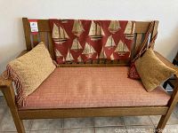 Full front view showing oak bench with padded seat, two beige tassel-trimmed pillows and red sailboat throw across backrest.