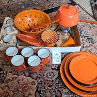 Photo showing full assortment of orange enamel kitchenware including cups, colander, spoon, funnel, saucepan with lid, utensil holder, and plates stacked separately.