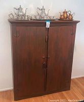 Closed front view of stained wooden armoire with vertical plank doors and metal latch; silver and copper tea sets on top (not included).