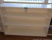 Photo showing front view of white wooden shelf unit with three shelves and wheels underneath.