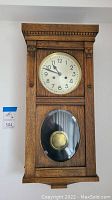 Front view of wooden wall clock mounted on wall, showing full clock face and pendulum window.