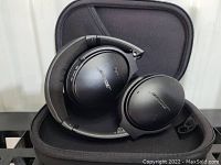 Black foldable Bose headphones with cushioned ear cups resting inside a black hard shell carrying case. USB charging cable visible.