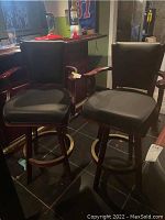 Pair of wooden barstools with black upholstery, showing the front view and bar setup background