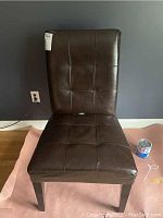 Brown leather desk chair with wooden legs from front, showing the smooth tufted upholstery on seat and backrest against a dark wall.