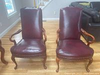 Front view of two burgundy leather armchairs with wooden arms and legs, showing metal rivet detailing.