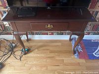 Front view of wooden console table showing dovetail drawer with brass drop handle and curved legs on wood floor.