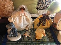 Photo shows five collectible dolls and figurines arranged on a patterned surface including bridal doll in white gown, Laines doll in yellow outfit, and three ceramic bird statues.