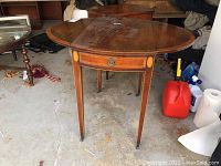 Full front view of the round drop leaf wooden side table with one leaf up. Shows drawer and tapered legs with casters.