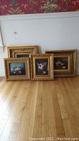 Four floral paintings with gold decorative frames shown leaning against a wall on wooden floor.